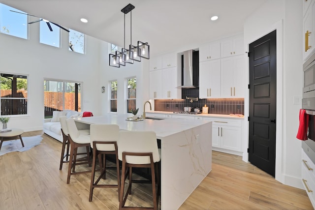 kitchen featuring light hardwood / wood-style flooring, a kitchen island with sink, white cabinetry, a kitchen bar, and wall chimney exhaust hood