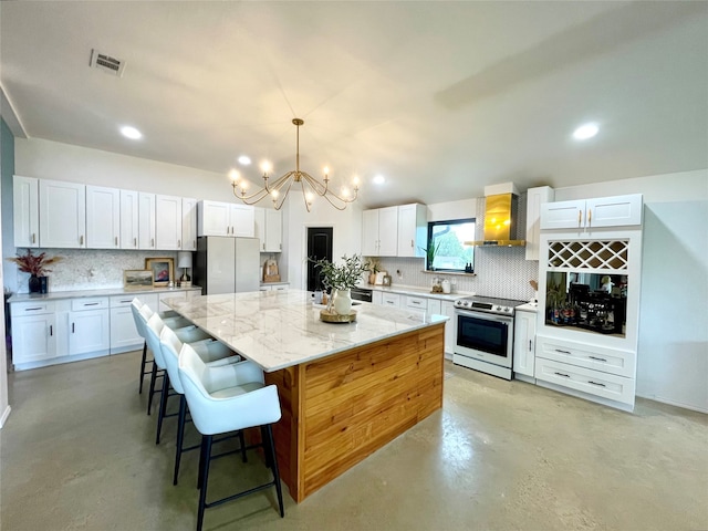 kitchen featuring stainless steel electric stove, refrigerator, wall chimney range hood, a kitchen island, and white cabinetry