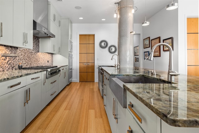 kitchen featuring dark stone counters, white cabinets, hanging light fixtures, wall chimney exhaust hood, and light wood-type flooring