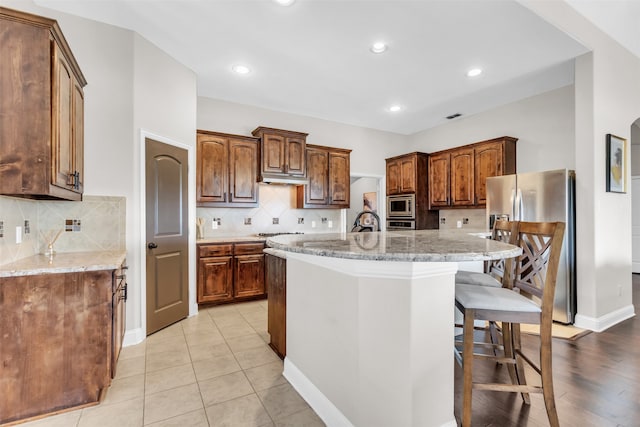 kitchen featuring a breakfast bar, a center island with sink, stainless steel appliances, light stone countertops, and backsplash