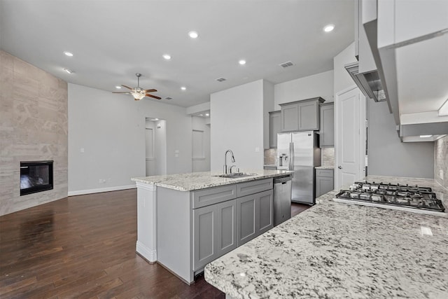 kitchen featuring a kitchen island with sink, gray cabinetry, a sink, appliances with stainless steel finishes, and a tiled fireplace