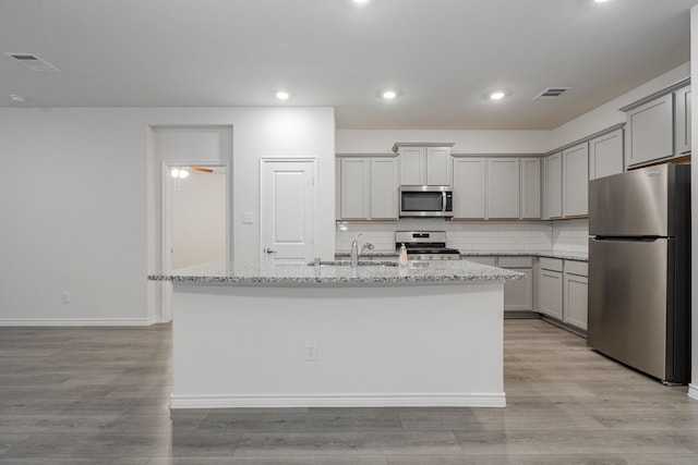 kitchen with gray cabinetry, light stone countertops, stainless steel appliances, and a kitchen island with sink