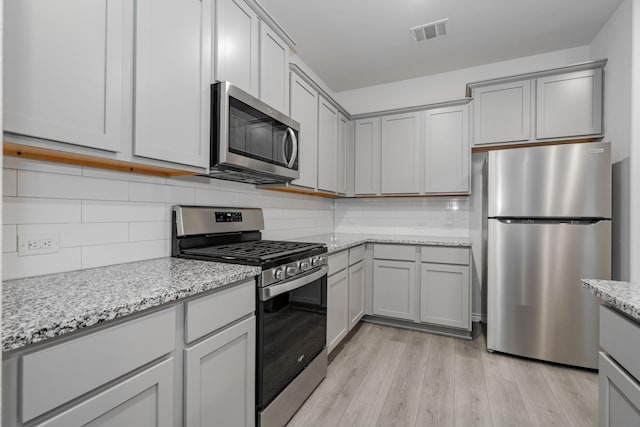 kitchen featuring gray cabinetry, stainless steel appliances, light stone counters, backsplash, and light wood-type flooring