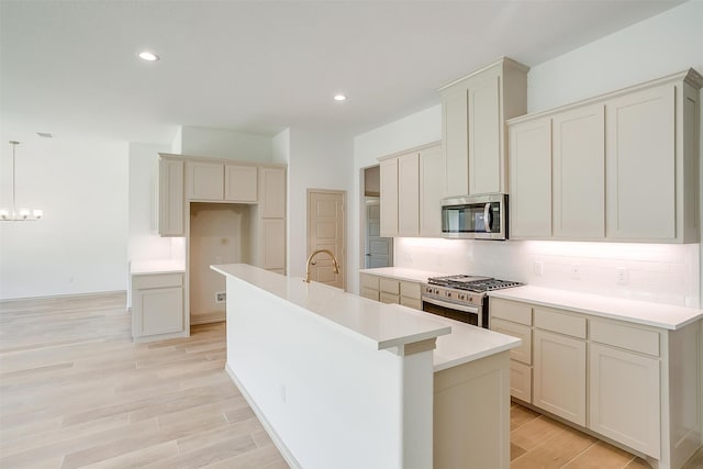 kitchen with hanging light fixtures, light wood-type flooring, an island with sink, appliances with stainless steel finishes, and a notable chandelier