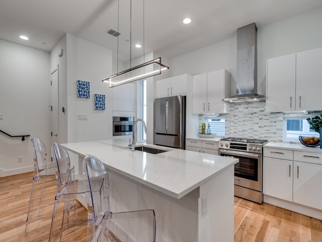 kitchen with wall chimney exhaust hood, stainless steel appliances, sink, hanging light fixtures, and an island with sink