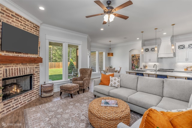 living room with a brick fireplace, ceiling fan, crown molding, sink, and hardwood / wood-style flooring