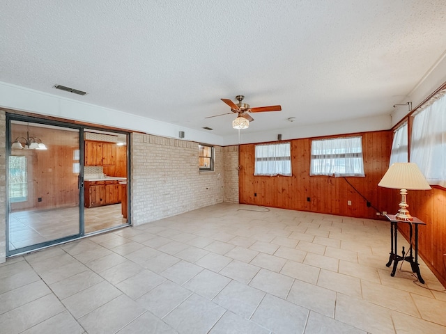 empty room featuring a textured ceiling, ceiling fan with notable chandelier, wooden walls, and brick wall