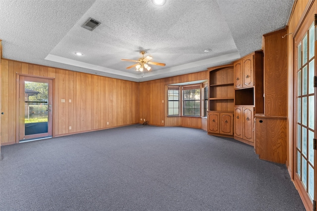 unfurnished living room featuring a tray ceiling, wood walls, ceiling fan, and dark carpet