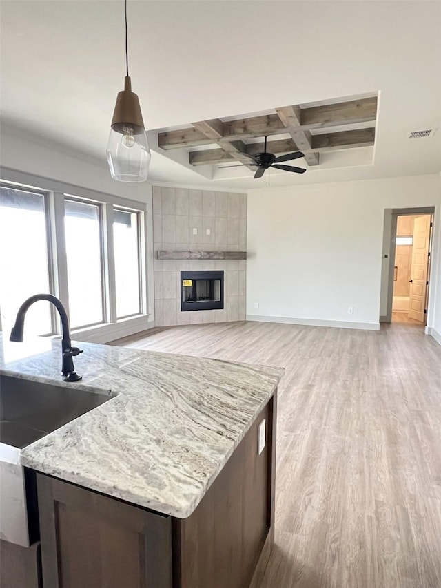 kitchen with coffered ceiling, light wood-type flooring, decorative light fixtures, a tile fireplace, and light stone countertops