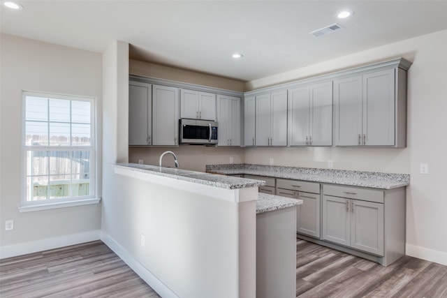 kitchen with gray cabinets, light wood-type flooring, light stone countertops, kitchen peninsula, and sink