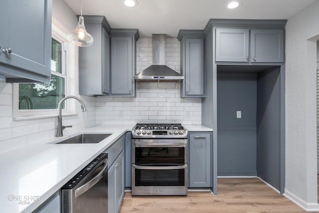 kitchen with sink, stainless steel appliances, wall chimney range hood, pendant lighting, and light hardwood / wood-style floors