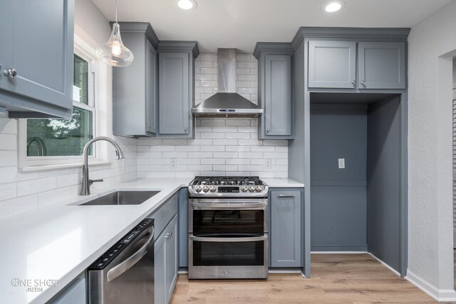 kitchen with sink, stainless steel appliances, wall chimney range hood, pendant lighting, and light hardwood / wood-style floors