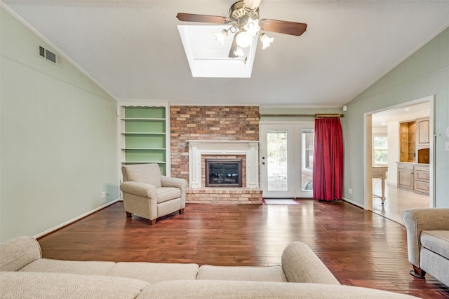 living room with built in shelves, ceiling fan, a brick fireplace, hardwood / wood-style floors, and vaulted ceiling with skylight