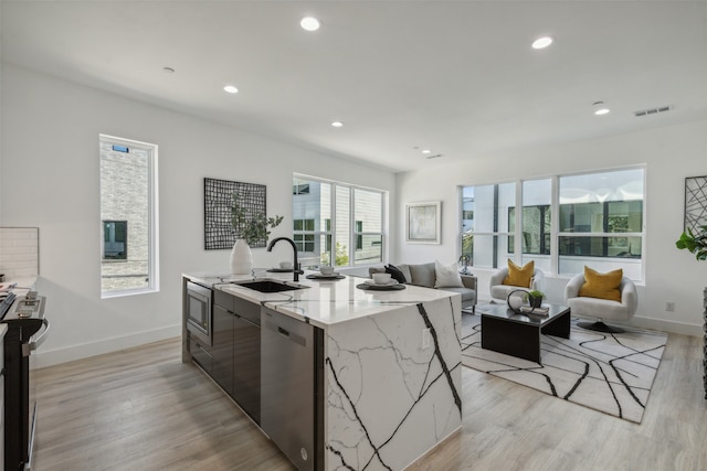 kitchen with light stone countertops, light wood-type flooring, stainless steel appliances, a kitchen island with sink, and sink