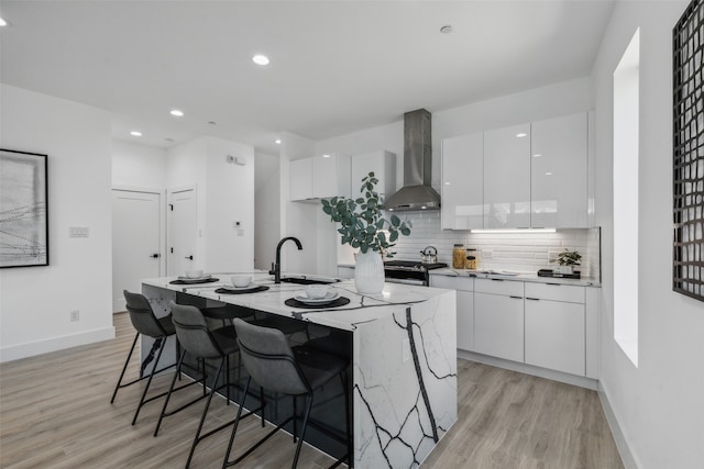 kitchen with white cabinets, wall chimney exhaust hood, light hardwood / wood-style floors, and an island with sink