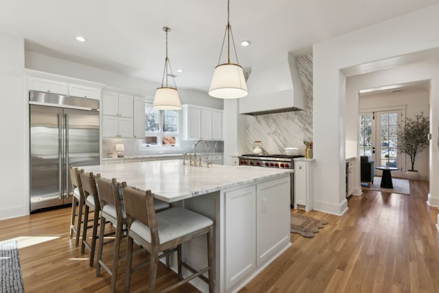 kitchen featuring a center island with sink, white cabinets, light wood-style flooring, stainless steel built in fridge, and custom exhaust hood