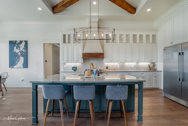 kitchen featuring stainless steel built in refrigerator, custom range hood, a kitchen island with sink, and beamed ceiling