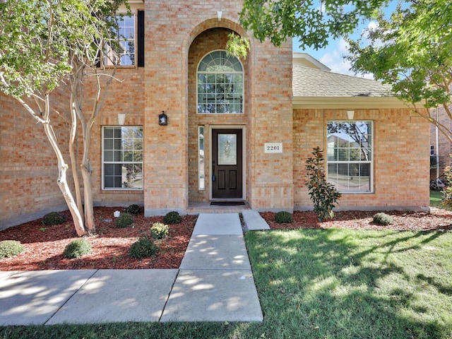 doorway to property featuring roof with shingles, a lawn, and brick siding