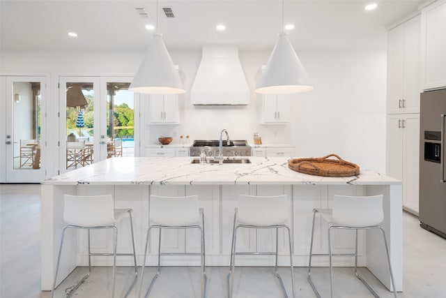 kitchen with a center island with sink, custom exhaust hood, stainless steel fridge, and light stone countertops