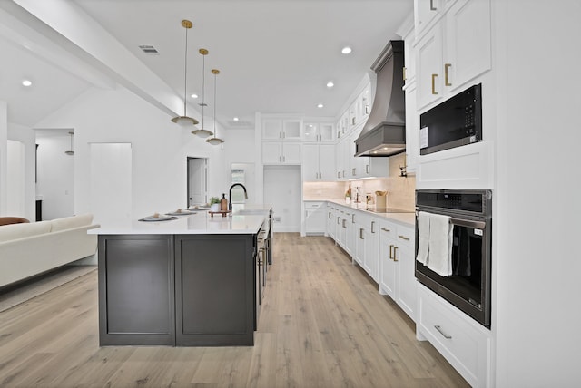 kitchen with black appliances, white cabinetry, custom range hood, and a kitchen island with sink