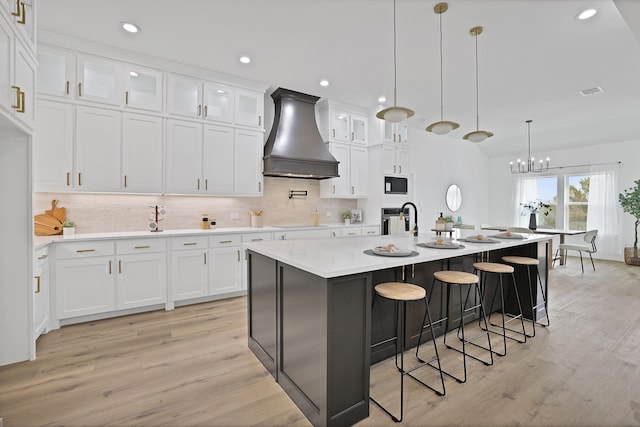 kitchen with premium range hood, pendant lighting, white cabinetry, oven, and an island with sink