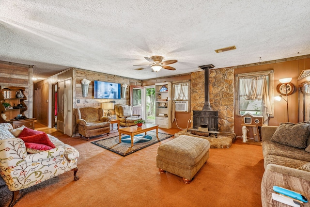 carpeted living room with ceiling fan, a wood stove, a textured ceiling, and wooden walls