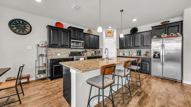 kitchen with a kitchen island with sink, sink, light wood-type flooring, light stone countertops, and stainless steel appliances