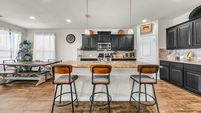 kitchen featuring backsplash, a kitchen island with sink, hanging light fixtures, appliances with stainless steel finishes, and light stone counters