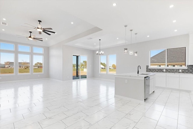 kitchen with a raised ceiling, decorative light fixtures, a center island with sink, white cabinets, and ceiling fan with notable chandelier