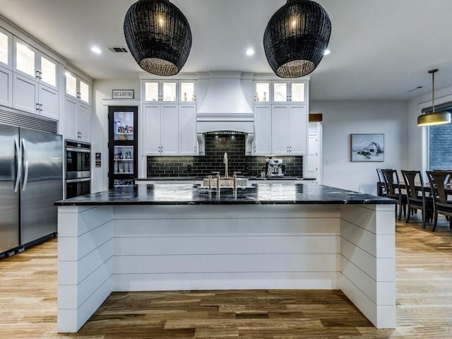 kitchen with custom exhaust hood, white cabinetry, decorative light fixtures, stainless steel appliances, and a kitchen island with sink