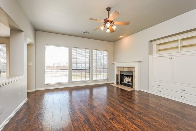 unfurnished living room with built in shelves, ceiling fan, dark wood-type flooring, and a tiled fireplace