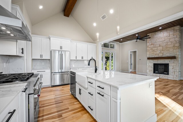 kitchen with wall chimney exhaust hood, white cabinetry, light stone counters, a center island with sink, and stainless steel appliances