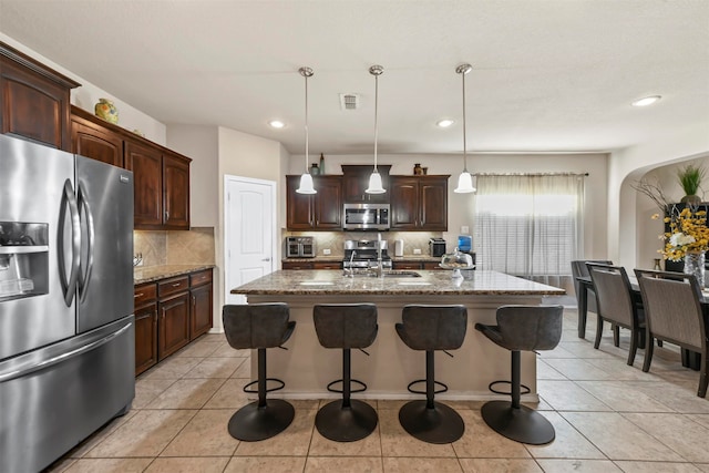 kitchen with pendant lighting, a kitchen island with sink, appliances with stainless steel finishes, light stone counters, and dark brown cabinetry