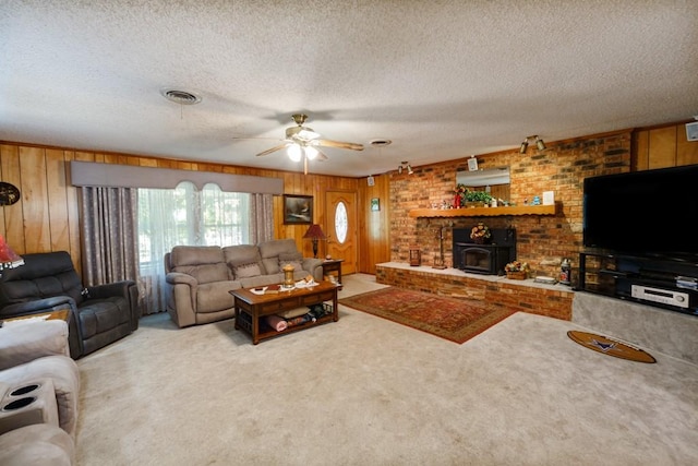 carpeted living room featuring a wood stove, wooden walls, ceiling fan, and a textured ceiling