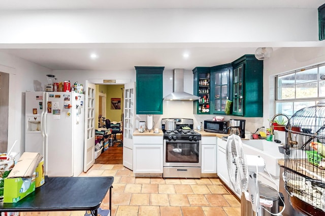 kitchen with blue cabinetry, white cabinetry, stainless steel gas stove, wall chimney exhaust hood, and white refrigerator with ice dispenser