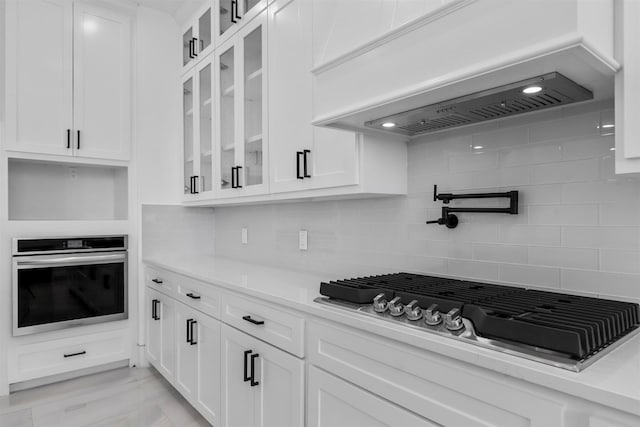 kitchen featuring gas cooktop, premium range hood, oven, decorative backsplash, and white cabinets