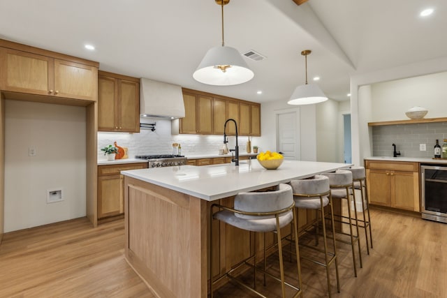 kitchen featuring premium range hood, a kitchen island with sink, decorative light fixtures, light hardwood / wood-style flooring, and wine cooler