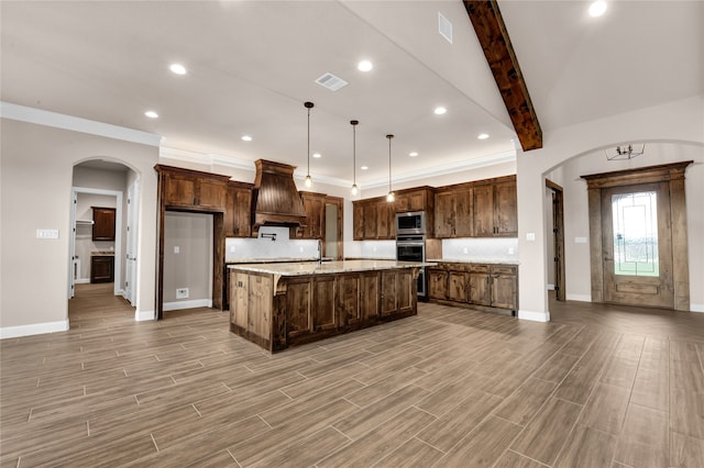 kitchen with premium range hood, light stone counters, beam ceiling, pendant lighting, and an island with sink