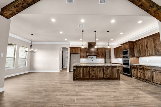 kitchen with custom exhaust hood, stainless steel appliances, a kitchen island with sink, a chandelier, and hanging light fixtures