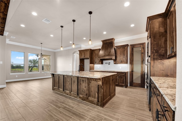 kitchen with a kitchen bar, premium range hood, light stone counters, a large island with sink, and hanging light fixtures