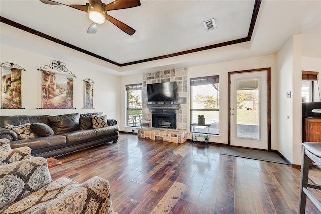 living room featuring dark hardwood / wood-style flooring, a raised ceiling, ceiling fan, crown molding, and a stone fireplace