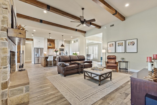 living room with beamed ceiling, ceiling fan, a stone fireplace, and light wood-type flooring