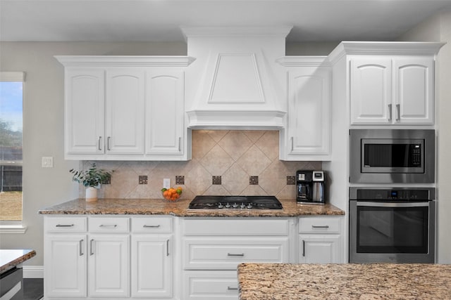 kitchen featuring white cabinets, custom range hood, stone counters, stainless steel appliances, and backsplash