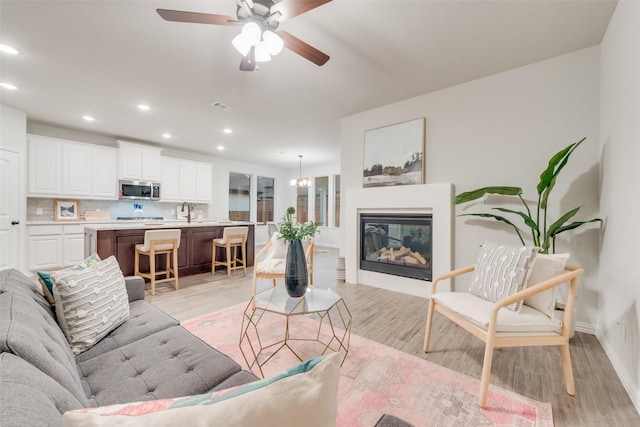 living area featuring visible vents, recessed lighting, light wood-style floors, a glass covered fireplace, and ceiling fan with notable chandelier