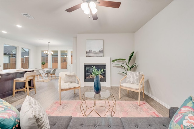 living room featuring visible vents, baseboards, light wood-style floors, a glass covered fireplace, and ceiling fan with notable chandelier