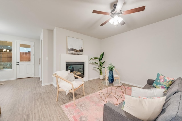 living room featuring a glass covered fireplace, light wood-style flooring, a ceiling fan, and baseboards