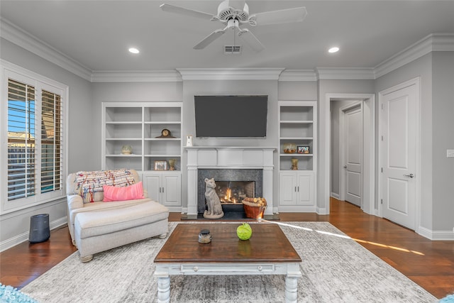 living room featuring ornamental molding, built in shelves, ceiling fan, a premium fireplace, and dark hardwood / wood-style floors