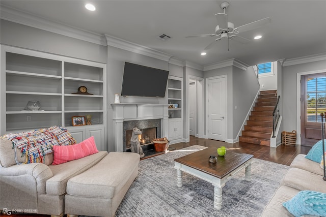 living room with built in shelves, ceiling fan, dark wood-type flooring, a high end fireplace, and crown molding