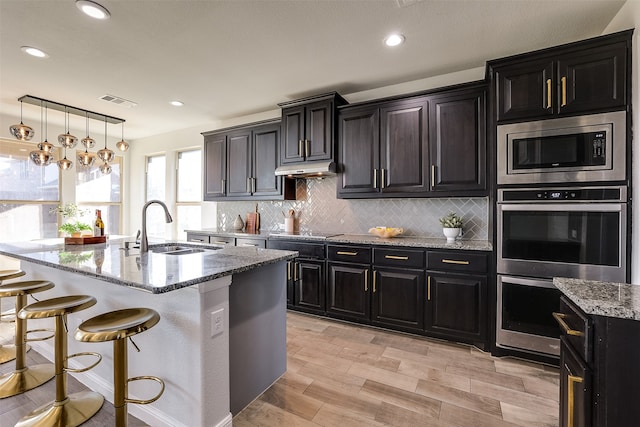 kitchen featuring sink, hanging light fixtures, light stone counters, an island with sink, and a breakfast bar area