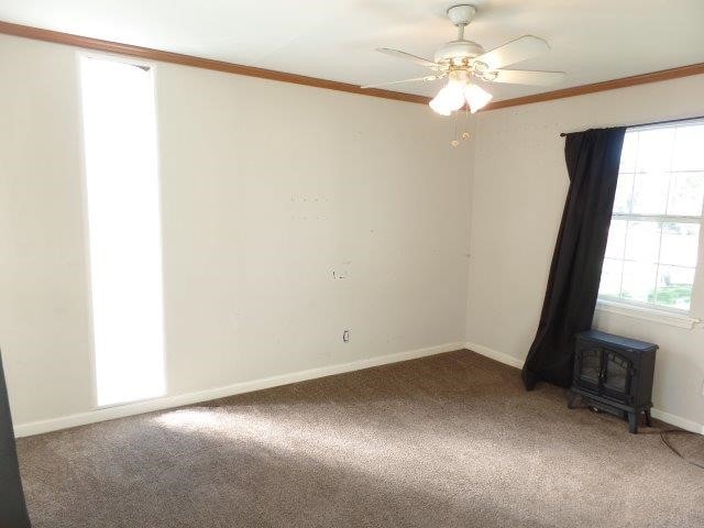 carpeted spare room with a wood stove, ceiling fan, and crown molding